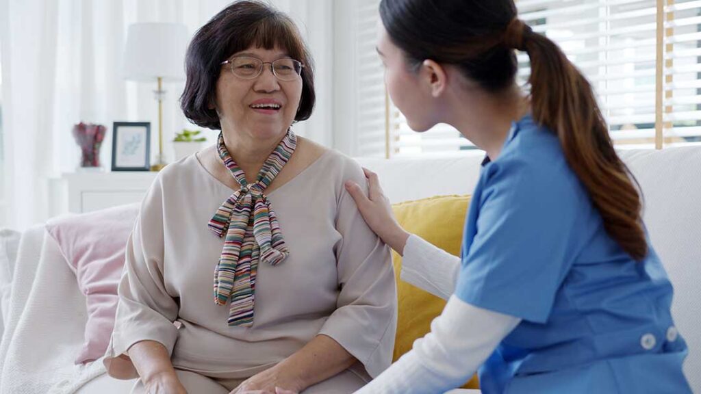 Older couple sharing a meal in an assisted living apartment while a caregiver provides discreet support.